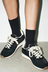 Zapatilla deportiva tipo running con diseño de paneles en contraste. Presenta cuerpo en color negro con refuerzos de gamuza sintética, detalle de pespuntes, talón en color crema y suela dentada bicolor.