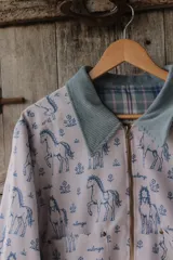 Campera de lona estampada con diseño de caballos y flores, con cuello de pana en color gris azulado y cierre frontal metálico. El interior está forrado en tela de viyela a cuadros.