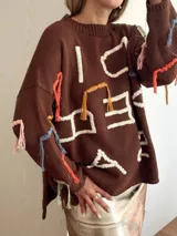 Sweater de punto color marrón con diseño oversize, cuello redondo y mangas largas. Presenta bordados artesanales en relieve con hilos de colores y detalles en lurex, incluyendo flecos decorativos distribuidos por toda la prenda.