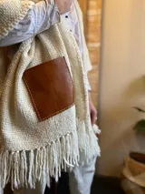 Ruana color crema tejida en lana merino, con flecos y bolsillos de cuero marrón.