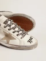 Zapatillas Golden Goose Super-Star de cuero blanco con efecto desgastado, estrella lateral y talón de cuero metalizado dorado, cordones con estampado animal print de leopardo, y detalles manuscritos en la puntera.