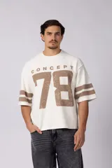 Remerón de corte oversize confeccionado en tejido rústico color crudo. Presenta un diseño de estilo deportivo con estampado frontal en color marrón que incluye el texto 'CONCEPT' y el número '78' con efecto desgastado. Posee mangas cortas con doble franja horizontal en color marrón.