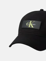 Gorra de béisbol negra de algodón tipo sarga, con visera curva y cierre ajustable con hebilla. Presenta un parche rectangular de silicona en el frente con el logo de Calvin Klein en color verde lima.