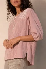 Blusa a rayas verticales en tonos rosados y blancos, con cuello redondo y cierre frontal parcial con botones. Presenta delicados detalles bordados en los hombros y mangas tres cuartos abullonadas con puños elásticos.