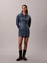 Vestido mini de denim azul con efecto desgastado, de manga larga, con cuello camisero, cierre frontal de botones metálicos y bolsillos con solapa en el pecho.