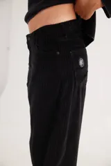 Pantalón de pana de corte ancho y tiro medio-bajo, con diseño de cinco bolsillos y acabado lavado. Presenta un aplique cuadrado decorativo en una de las piernas y un pin bordado de la marca.