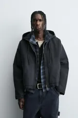 Cazadora tipo bomber de denim color negro con efecto lavado, corte boxy fit, cierre frontal de cremallera, capucha ajustable con cordón y bolsillos laterales de vivo.
