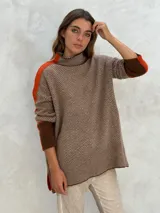 Sweater de punto con patrón de espiga en tonos beige y marrón, cuello alto, mangas largas con detalles en naranja y puños marrones.