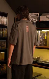 Remera de manga corta con cuello redondo y calce oversize, color gris oscuro. Presenta un estampado frontal con estética retro que incluye figuras humanas y una computadora antigua en tonos rojos.