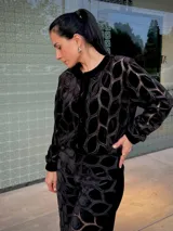 Chaqueta tipo bomber color negro con diseño de patrón geométrico calado y textura aterciopelada. Presenta cuello redondo, cierre frontal con botones y puños elásticos.