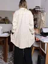 Sobre camisa color manteca con costuras decorativas en zigzag de color oscuro. Presenta cuello clásico, cierre frontal con botones metálicos a presión, mangas largas con puños abotonados y un bolsillo frontal con solapa.