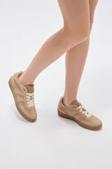 Zapatillas urbanas de corte bajo en tonos beige y tostado, combinando paneles de cuerina y gamuza sintética. Cuentan con cordones anchos tipo cinta de raso en color crudo, pespuntes decorativos y suela de goma al tono.