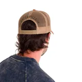 Gorra tipo trucker con frente y visera en color verde oliva y marrón, con panel trasero de malla en color beige. Presenta un parche circular bordado en el frente con el texto 'A Tractor Quality Company' y cierre ajustable snapback.