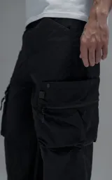 Pantalón cargo oversize de corte relajado, confeccionado en textil técnico resistente de color negro. Presenta bolsillos laterales cargo de gran tamaño con cierres metálicos, bolsillos laterales superiores y bolsillos traseros con broches a presión. Incluye cinturón integrado de cinta de seguridad con hebilla de encastre técnica.