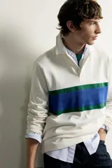 Remera polo de manga larga de corte regular fit, confeccionada en algodón color blanco crudo. Presenta cuello solapa blanco, cierre frontal con botonadura oculta y una franja horizontal ancha en el pecho en color azul y verde.