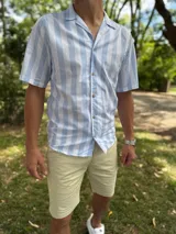 Camisa de manga corta con diseño a rayas verticales en color celeste y blanco, cuello abierto estilo cubano y cierre frontal con botones marrón claro.