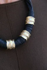 Collar de diseño artesanal compuesto por múltiples hilos de cordón negro entrelazados, intercalados con piezas cilíndricas de metal dorado con textura irregular y acabado rústico.
