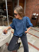 Blusa de denim liviano de corte asimétrico en tono azul, con mangas abullonadas, cuello redondo y cierre lateral con lazos para atar y botones.