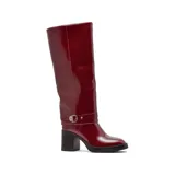 Bota de caña alta Korium modelo Sira, confeccionada en cuero sintético negro con acabado brillante. Presenta un taco cuadrado robusto, punta redondeada y una tira decorativa con hebilla metálica en la zona del tobillo.