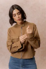 Chaqueta de gamuza sintética color tostado, con cuello mao, cierre frontal con cremallera y bolsillos frontales con solapa. Presenta detalles de broches metálicos y puños ajustables.