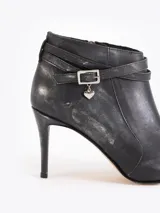 Bota de caña baja color negro con efecto gastado, confeccionada en cuero vacuno. Presenta punta fina, taco fino, y tira con hebilla y dije de corazón colgante alrededor del tobillo.