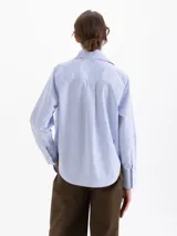 Camisa de poplin color blanco, con cuello clásico, cierre frontal de botones y mangas largas con puños. Presenta un diseño bordado de pequeñas llamas distribuidas por toda la prenda.