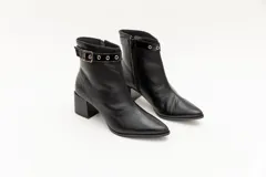 Botas de cuero negro, de caña baja, punta fina y taco cuadrado. Presentan un aplique charolado al tono, una correa con hebilla y ojales metálicos.