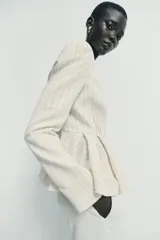 Blazer entallado de color crema con rayas verticales finas, cuello sin solapa y manga larga con hombreras. Presenta un diseño con bajo en evasé tipo peplum y cierre frontal con botones negros.
