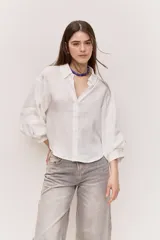 Camisa blanca de corte amplio y largo cropped, con mangas abullonadas, puño ajustado, cuello clásico y abertura en V con botones al tono.