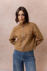 Chaqueta de gamuza sintética color tostado, con cuello mao, cierre frontal con cremallera y bolsillos frontales con solapa. Presenta detalles de broches metálicos y puños ajustables.