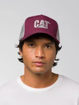 Gorra tipo trucker color violeta con panel frontal de sarga y paneles traseros de malla gris. Presenta el logo de Caterpillar bordado en la parte frontal y visera curva.