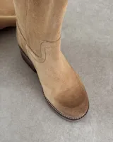 Botín corto de cuero vacuno color beige, estilo cowboy, con caña media y costuras decorativas en forma de onda en el empeine. Presenta un taco bajo y suela con pespunte visible.