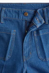 Pantalón de jean de tiro alto, color azul medio, con pierna ancha y corte ajustado en la cadera. Presenta bolsillos de parche tanto en la parte delantera como en la trasera.