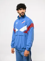 Buzo deportivo de nylon con medio cierre frontal y cuello alto. Presenta un diseño de bloques de color en azul, blanco y bordó, con el escudo del Club Nacional de Football y el logo de Umbro en el pecho.
