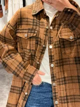 Camisa tipo sobrecamisa de corte oversize, confeccionada en tela a cuadros en tonos marrones y negros. Presenta cierre frontal con botones metálicos a presión, bolsillos frontales con solapa y mangas largas con puños abotonados.