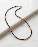Collar de diseño minimalista compuesto por pequeñas cuentas negras intercaladas con cuentas cilíndricas de piedra natural en tono coral y detalles en dorado.