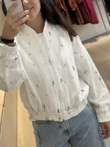 Campera tipo bomber color blanco con estampado de motivos deportivos, cierre frontal completo, cuello redondo y puños elásticos.