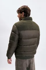 Campera puffer color verde oliva oscuro con diseño capitoneado. Presenta un panel superior en tejido tipo corderito en el pecho y cuello alto. Cuenta con cierre frontal completo, puños y cintura elástica acanalada, y un bolsillo vertical con cierre en el pecho.
