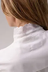 Camisa de algodón color gris azulado, con cuello alto, cierre frontal de botones y mangas largas con puños abotonados. Presenta un diseño con pliegues y detalles de encaje blanco en forma de V en el frente.