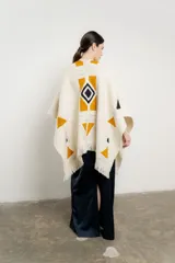 Ruana de tejido grueso color crema con flecos en el borde inferior y diseño geométrico en ocre, negro y gris.