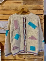 Cardigan de lana merino tejido a mano con técnica de dos agujas, de color marfil con parches geométricos de tela en tonos celestes y lila aplicados con costuras visibles. Presenta cierre frontal con botones de madera y detalles de ribetes multicolores en los bordes.