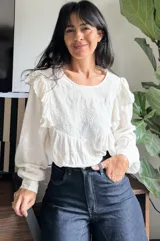 Camisa blanca de manga larga con detalles de volados en los hombros y bordado geométrico en el pecho. Presenta un diseño holgado con puños elásticos.