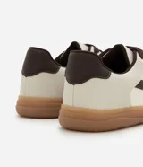 Championes de estilo urbano con diseño retro, confeccionados en material sintético color crema con detalles en marrón oscuro. Presentan recortes laterales, cierre con cordones y suela de goma color caramelo.