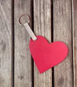 Llavero con forma de corazón de cuero rojo con textura granulada, tira de cuero color beige y aro metálico plateado.