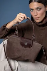 Cartera pequeña de mano confeccionada en gamuza color marrón, con solapa de cuero liso y cierre metálico. Incluye correa larga ajustable y extraíble para usar al hombro o cruzada.