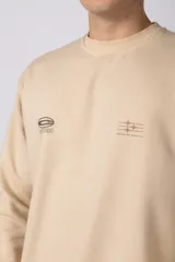 Buzo de cuello redondo en color arena, con diseño de calce regular y estampado gráfico minimalista en el pecho. Presenta puños y ruedo acanalados, con un pequeño detalle de etiqueta en el puño izquierdo.