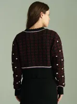 Cardigan de punto negro y rojo con diseño texturizado, cuello redondo y cierre frontal con botones. Presenta detalles de ribetes en blanco y violeta en el cuello, la botontera, los puños y el ruedo, que cuenta con flecos colgantes.
