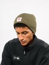 Gorro tipo beanie de tejido de punto acanalado en color verde oliva, con parche frontal rectangular blanco que presenta el logo de Umbro en rojo.