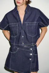 Camisa corta de denim de manga corta, con cuello solapa, escote en V y bolsillos delanteros de plastrón. Cuenta con costuras en contraste y cierre frontal con botones metálicos.