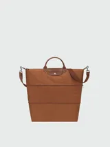 Bolso de viaje Longchamp modelo Le Pliage Original, de diseño ampliable, confeccionado en nylon negro con detalles en cuero marrón en las asas y solapa frontal con cierre de botón a presión. Incluye correa ajustable y extraíble.